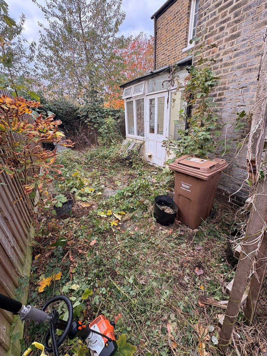 Partly cleared garden beside the house in Ware, with cut growth on the ground, a brown garden waste bin and a strimmer in the foreground