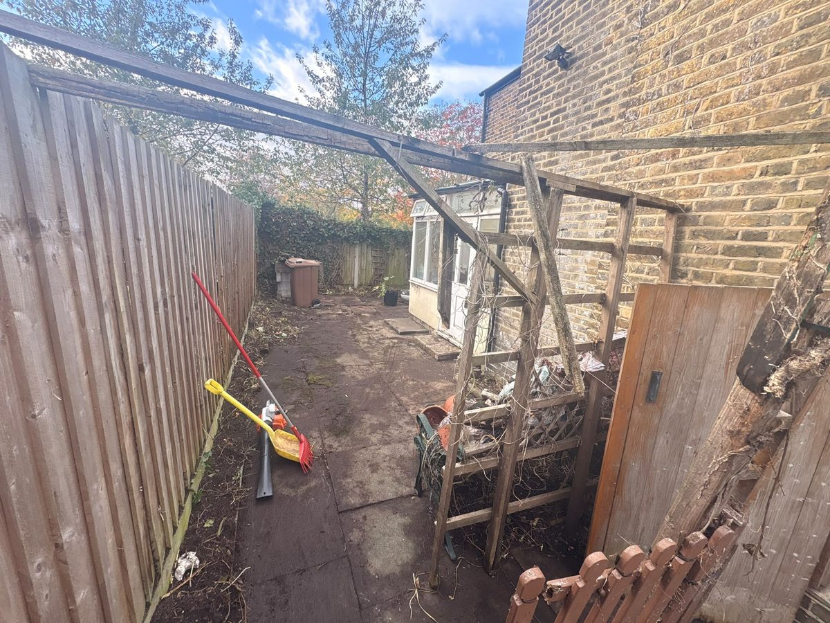 Cleared narrow side passage beside a close-boarded fence in Ware, with an old timber frame overhead and a gate at the end