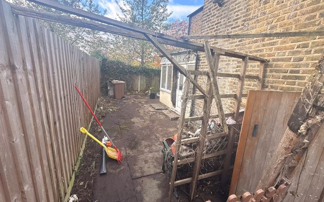 Cleared narrow side passage beside a close-boarded fence in Ware, with an old timber frame overhead and a gate at the end